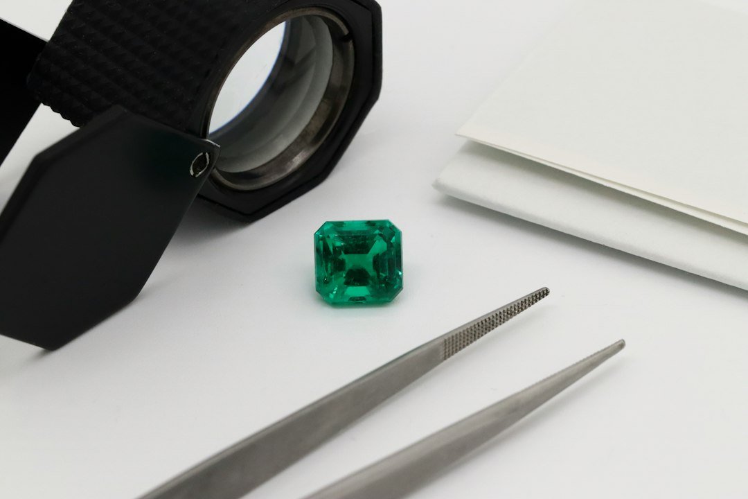 Cut emerald gemstone beside a jeweller's loupe and tweezers on a white surface